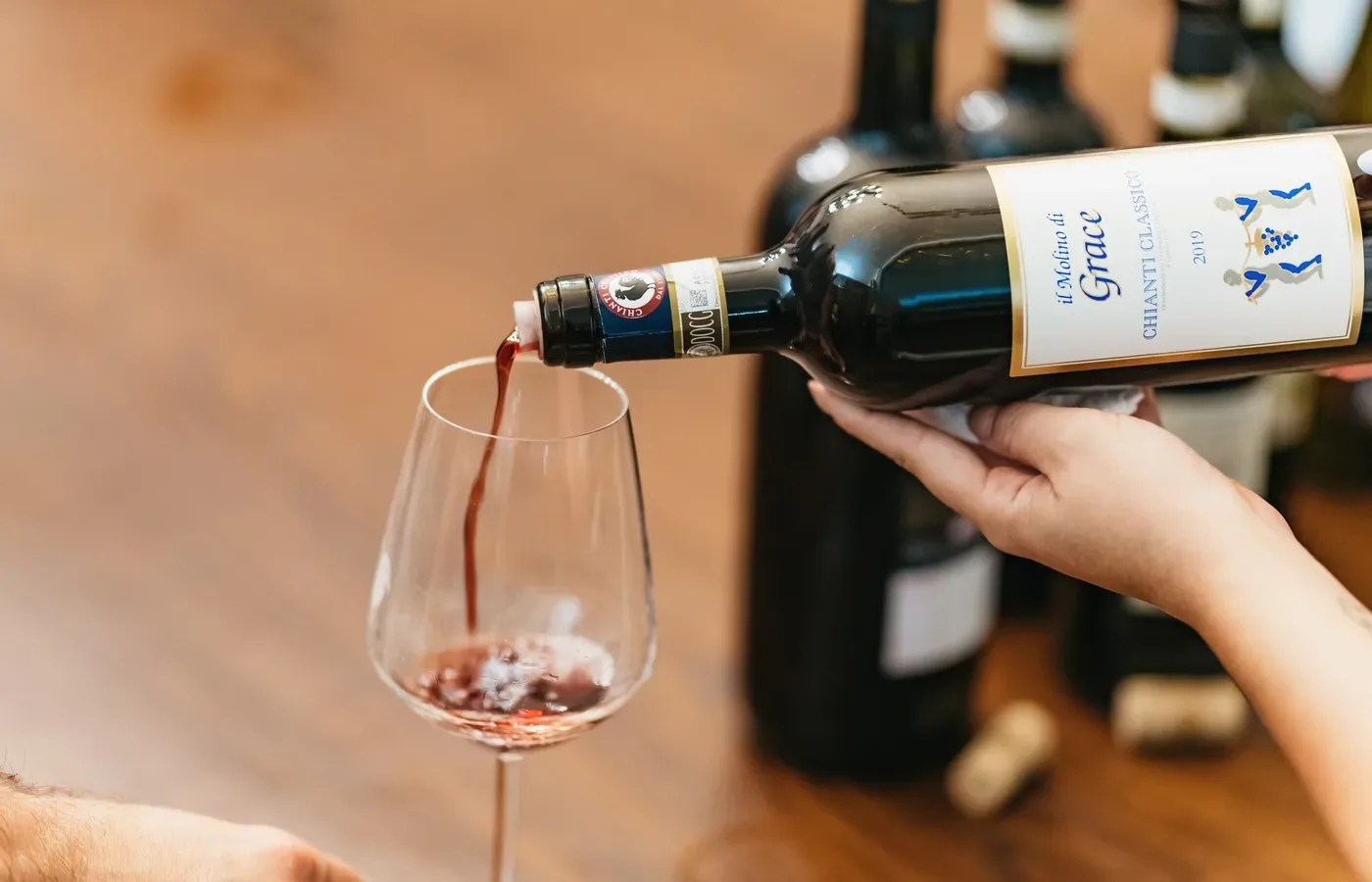 A bottle of Chianti Classico being poured into a tasting glass at a winery, with the Gallo Nero black rooster seal of the Consorzio Chianti Classico and the gold DOCG band both visible on the bottle neck.