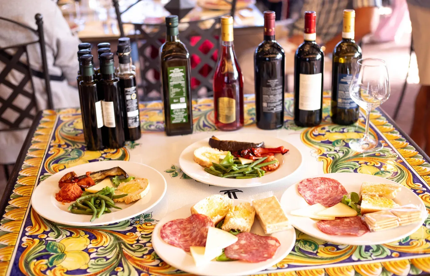 A tasting laid out on a painted majolica table at a Chianti winery: plates of sliced salami, wedges of pecorino cheese, bruschetta and crostini, grilled peppers and aubergine, beside bottles of red, rosé and white wine and dark bottles of olive oil, with an empty tasting glass.